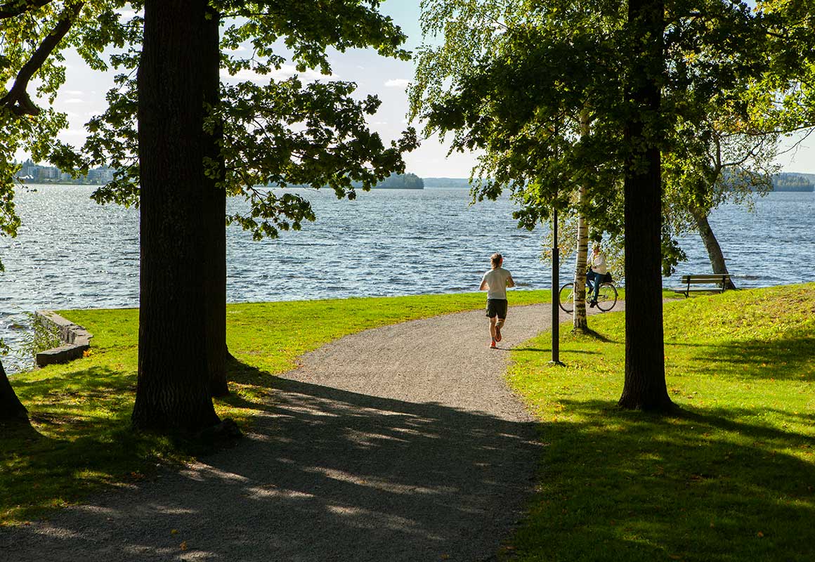 Tampereen Härmälänrannan puistossa on polku veden äärellä. Juokseva henkilö ja pyöräilevä henkilö näkyvät puiden varjossa järven rannalla.